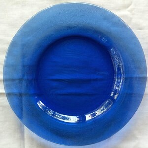 May include: A circular, translucent cobalt blue glass plate with a smooth, polished surface and a wide, flat rim. The plate features a deep, saturated blue hue that allows light to pass through, highlighting the uniform glass construction.