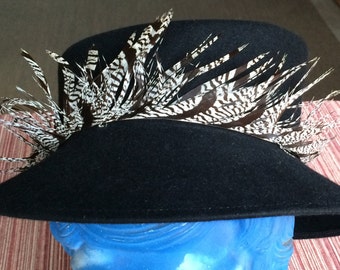 Puede incluir: Un sombrero de fieltro negro con ala ancha y adorno de plumas. Las plumas son negras y blancas con un patrón a rayas.