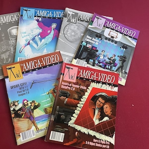 May include: A collection of vintage "AMIGA-VIDEO Journal" magazines. The covers feature colorful illustrations and text, including "Update City!", "Return of Toaster Tricks", and "Video Toaster & Lightwave 3D". The magazines are arranged on a red surface.