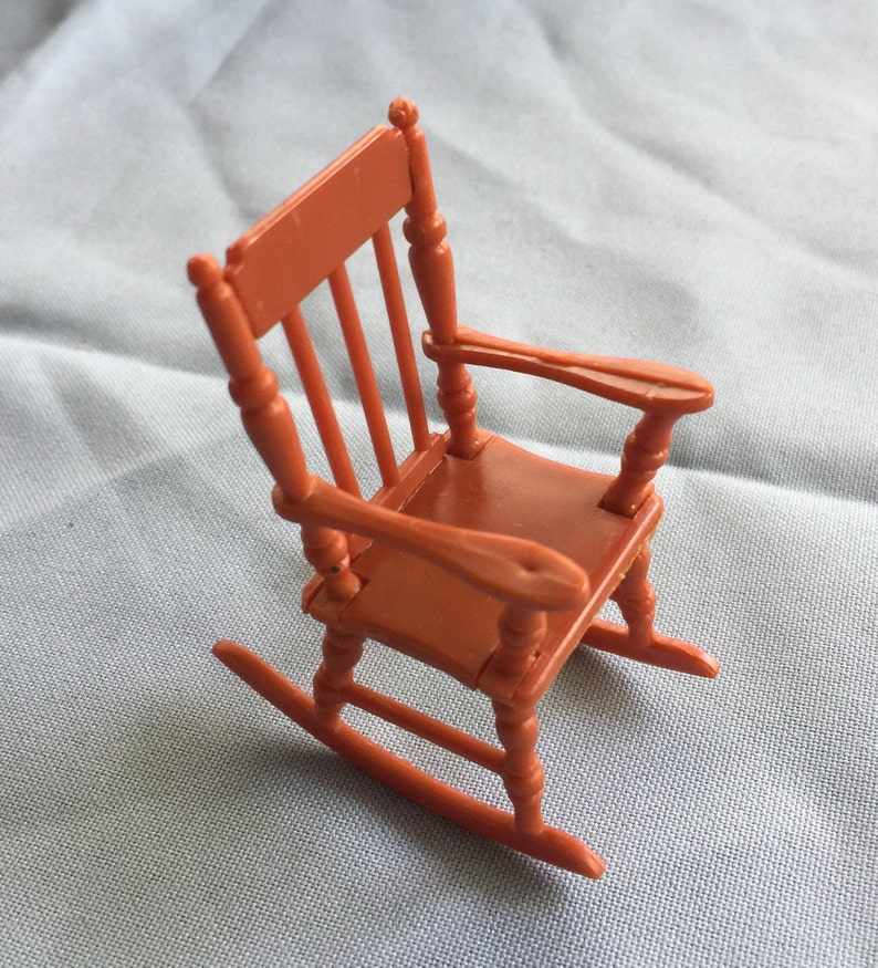 K&ouml;nnte beinhalten: Ein Miniatur-Schaukelstuhl aus Plastik, in rotbrauner Farbe lackiert. Der Stuhl hat ein einfaches Design mit einer geschwungenen R&uuml;ckenlehne und einer breiten Sitzfl&auml;che.