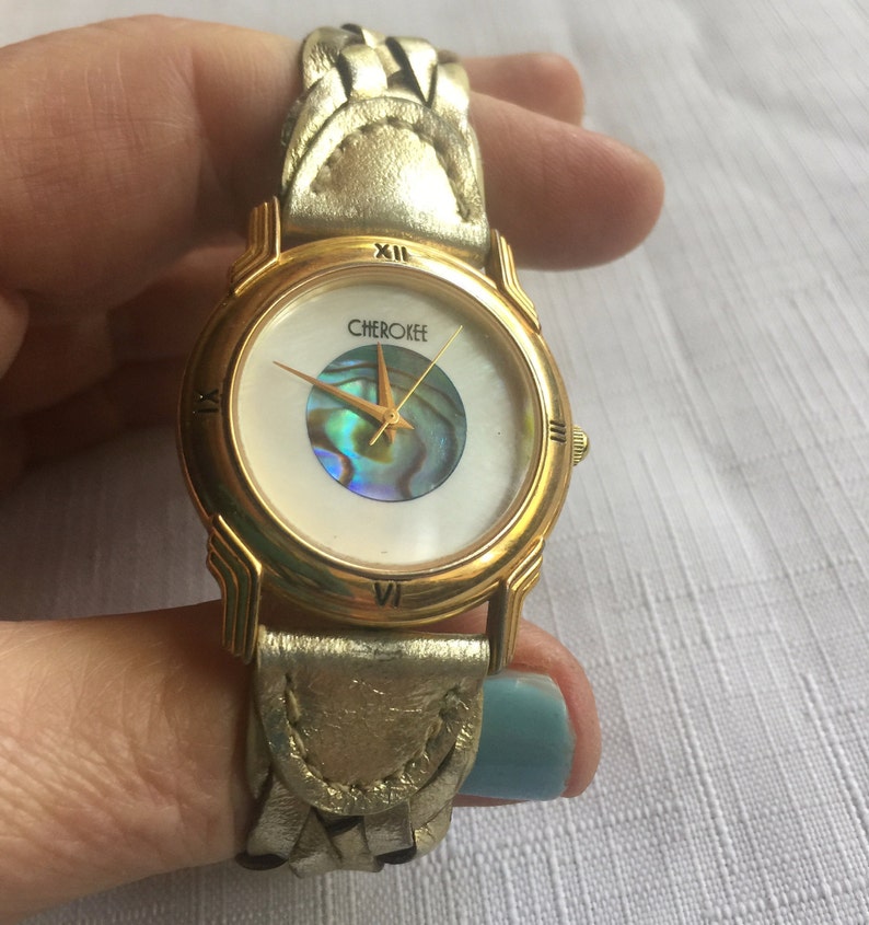 May include: Gold-tone wristwatch with a white face and a mother-of-pearl dial. The watch has a gold-tone band with a braided leather design. The watch face says "Cherokee" in gold lettering.