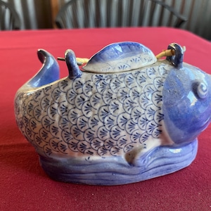 Puede incluir: Tetera de cerámica con forma de pez, con un diseño de escamas azules y blancas. La tetera tiene tapa y asa, y está sobre una superficie roja. Es un artículo decorativo.