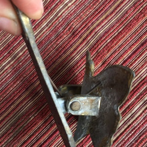 May include: A vintage brass-toned paper clip with a decorative leaf design. The clip has a hinged mechanism and a pointed end for inserting into paper. The clip is held against a red and tan striped background.