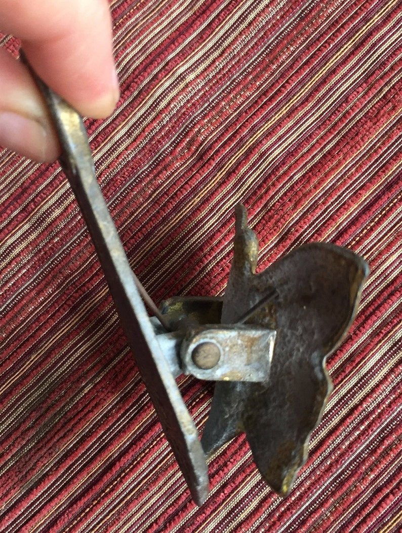 May include: A vintage brass-toned paper clip with a decorative leaf design. The clip has a hinged mechanism and a pointed end for inserting into paper. The clip is held against a red and tan striped background.