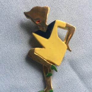 Puede incluir: Un broche dorado que representa una figura estilizada con un vestido amarillo y zapatos verdes. La figura tiene un corpi&ntilde;o negro y azul.