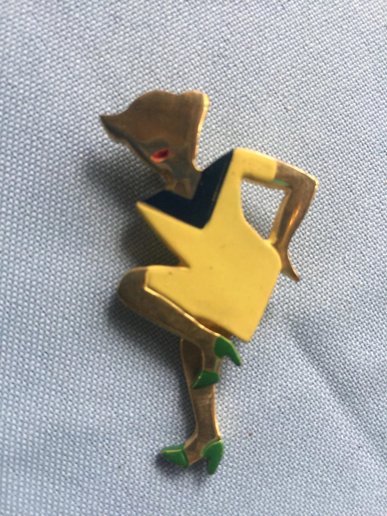 Puede incluir: Un broche dorado que representa una figura estilizada con un vestido amarillo y zapatos verdes. La figura tiene un corpi&ntilde;o negro y azul.