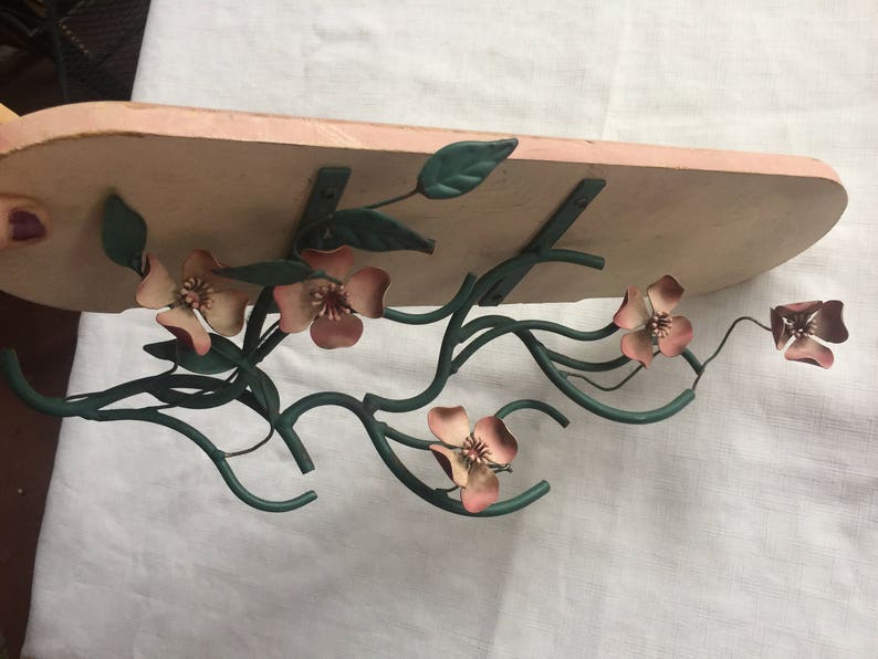 May include: A metal shelf with a pink painted wood base. The shelf is decorated with a metal floral design with pink flowers and green leaves.