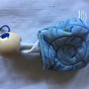 May include: A blue and white fabric snail with a wooden head and blue eyes. The snail is made of soft fabric and is suitable for children over 36 months.