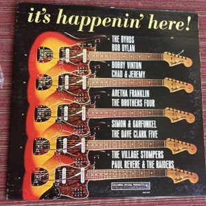 May include: A vintage record album cover with a black background and a colorful image of six electric guitars. The album title is "it's happenin' here!" and features a list of artists including The Byrds, Bob Dylan, Bobby Vinton, Chad & Jeremy, Aretha Franklin, The Brothers Four, Simon & Garfunkel, The Dave Clark Five, The Village Stompers, and Paul Revere & The Raiders.