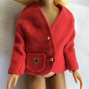 May include: A doll wearing a bright red, textured, long-sleeved jacket with a V-neckline. The jacket features two small, round, gold-colored buttons down the front and a single patch pocket on the right side adorned with a small, metallic, shield-shaped emblem.