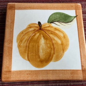 May include: A ceramic tile with a brown frame and a white background. The tile features a hand-painted image of a yellow pumpkin with a green leaf.