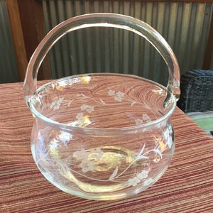 May include: A clear glass bowl with a floral design etched into the glass. The bowl has a handle and is perfect for serving snacks or fruit.