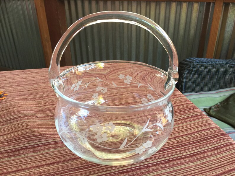 May include: A clear glass bowl with a floral design etched into the glass. The bowl has a handle and is perfect for serving snacks or fruit.
