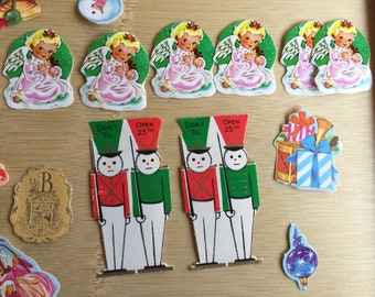 以下が含まれることがあります： ヴィンテージのクリスマスをテーマにした紙の切り抜きコレクション。切り抜きには、ピンクのドレスを着た天使、テキスト「Don't Til 25th」の兵隊、ギフトボックス、装飾オーナメントが含まれています。カラーパレットは主に赤、緑、白です。