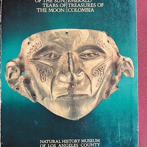 Puede incluir: Una máscara de oro con detalles intrincados, que representa un rostro humano. La máscara se muestra sobre un fondo azul verdoso con el texto "SWEAT GOLD AND OF THE SUN, EMERALD TEARS OF TREASURES OF THE MOON: COLOMBIA" y "NATURAL HISTORY MUSEUM OF LOS ANGELES COUNTY".