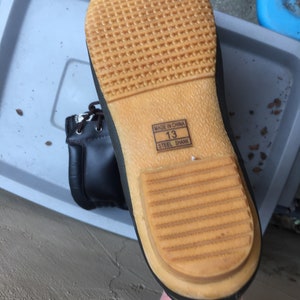 May include: Close-up of the bottom of a black leather boot with a brown rubber sole. The sole has a textured pattern and the words "Made in China" and "Steel Shank" are printed on the side. The boot size is "13".