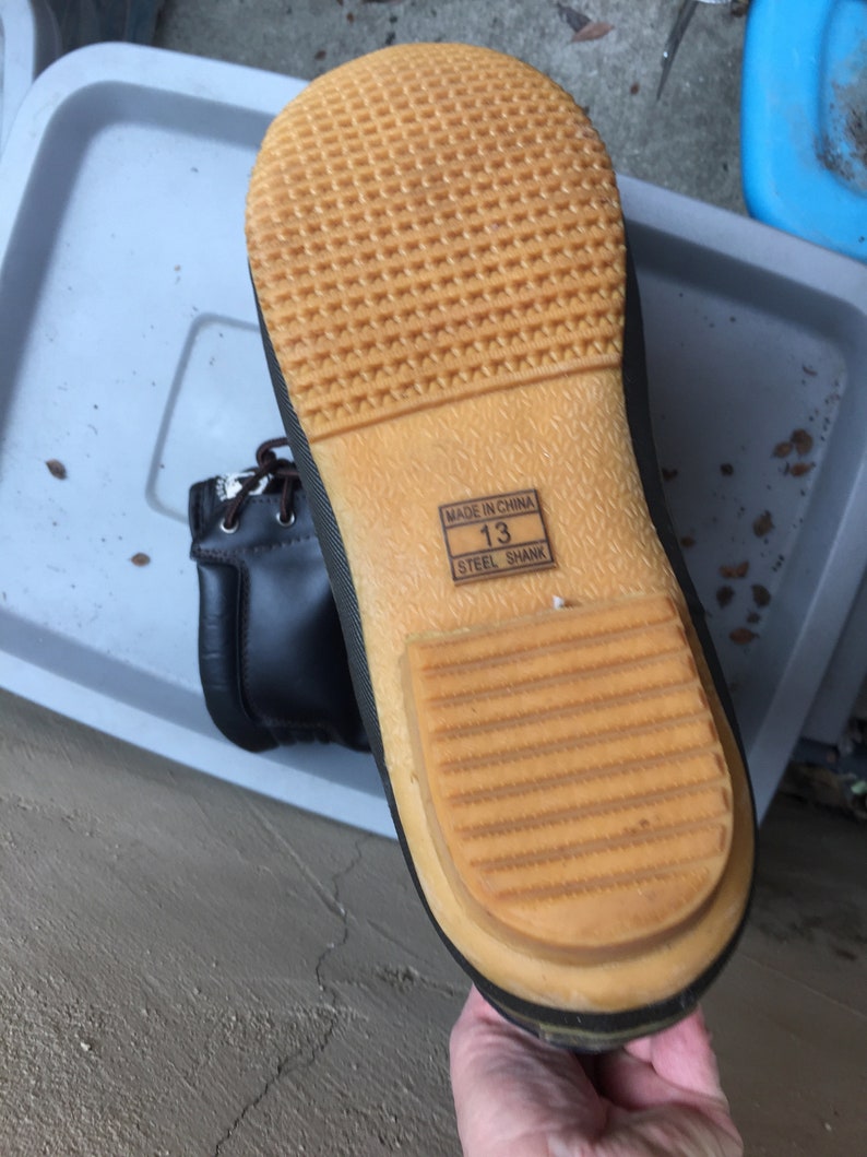 May include: Close-up of the bottom of a black leather boot with a brown rubber sole. The sole has a textured pattern and the words "Made in China" and "Steel Shank" are printed on the side. The boot size is "13".