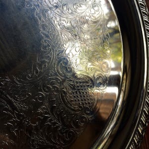 May include: A silver serving tray with an ornate, embossed design. The tray has a raised rim and a smooth, reflective surface.