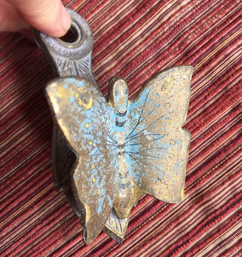 May include: A vintage metal butterfly-shaped object with a handle. The butterfly has blue and gold paint with black details. The handle is a light gray color with decorative detailing.