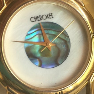 May include: A gold-tone wristwatch with a white face and a mother-of-pearl dial. The watch has Roman numeral hour markers and a gold-tone band. The watch face features a round, iridescent abalone shell design in the center and the word "CHEROKEE" printed above it.