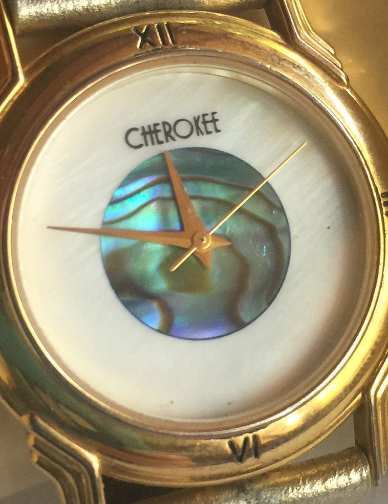 May include: A gold-tone wristwatch with a white face and a mother-of-pearl dial. The watch has Roman numeral hour markers and a gold-tone band. The watch face features a round, iridescent abalone shell design in the center and the word "CHEROKEE" printed above it.