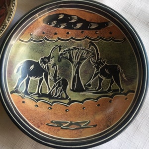 May include: A round, black slate serving dish with a carved design of three elephants and a tree. The elephants are black and the tree is green. The background is a light brown color.