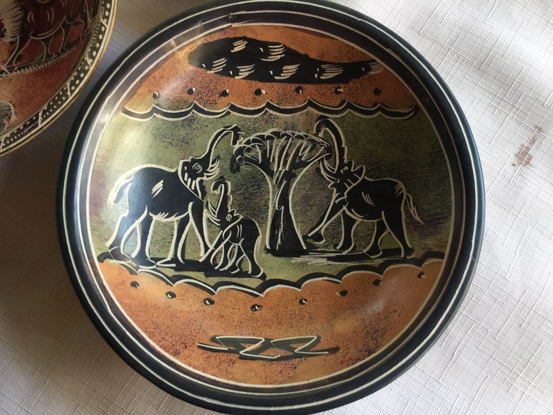 May include: A round, black slate serving dish with a carved design of three elephants and a tree. The elephants are black and the tree is green. The background is a light brown color.