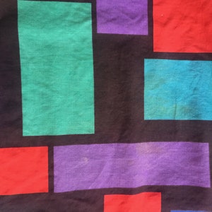K&ouml;nnte beinhalten: Ein schwarzer Stoff mit einem geometrischen Muster aus bunten Quadraten. Die Quadrate sind in den Farben Rot, Lila, Gr&uuml;n, Blau und Gelb.