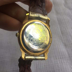 May include: Gold-tone watch with a brown leather braided band. The watch face is covered with a clear glass. The back of the watch has the words "Cherokee" and "Water Resistant" printed on it.