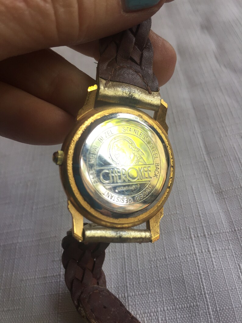 May include: Gold-tone watch with a brown leather braided band. The watch face is covered with a clear glass. The back of the watch has the words "Cherokee" and "Water Resistant" printed on it.