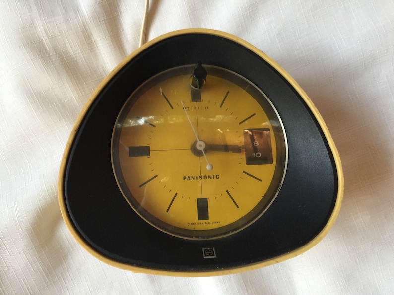 May include: A yellow and black alarm clock with a round face and black hands. The clock face is yellow and has the word "Panasonic" printed on it. The clock is made in Japan.
