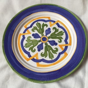 May include: A white ceramic plate with a blue, green and yellow floral design. The plate has a wide rim with a blue and green border.