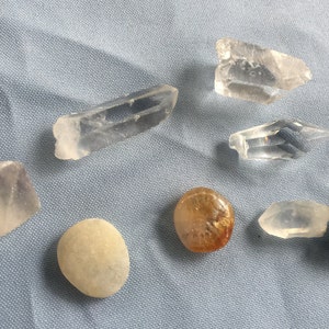 May include: A collection of seven crystals and stones, including clear quartz, amethyst, a golden rutilated quartz, and a black stone. The crystals and stones are arranged on a blue fabric background.