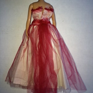 May include: A doll wearing a formal gown. The dress features a cream-colored bodice and skirt, overlaid with a red tulle fabric. The tulle cascades down the back, creating a flowing effect. The doll has blonde hair.