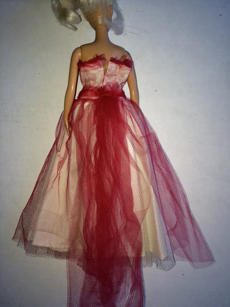 May include: A doll wearing a formal gown. The dress features a cream-colored bodice and skirt, overlaid with a red tulle fabric. The tulle cascades down the back, creating a flowing effect. The doll has blonde hair.