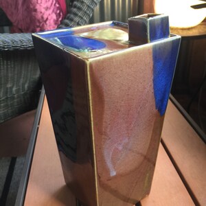 Op de afbeelding: Een hoge, rechthoekige keramische vaas met een bruine, blauwe en gouden glazuur. De vaas heeft een vierkante opening aan de bovenkant en een uniek, abstract ontwerp.
