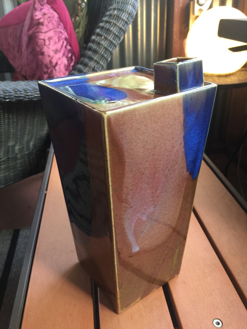 Op de afbeelding: Een hoge, rechthoekige keramische vaas met een bruine, blauwe en gouden glazuur. De vaas heeft een vierkante opening aan de bovenkant en een uniek, abstract ontwerp.