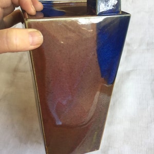 Op de afbeelding: Een keramische vaas met een vierkante basis en een rechthoekig lichaam. De vaas is geglazuurd in tinten bruin, blauw en paars. De bovenkant van de vaas heeft een vierkant deksel.