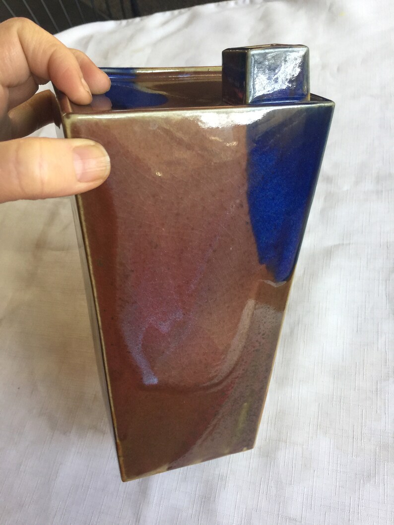 Op de afbeelding: Een keramische vaas met een vierkante basis en een rechthoekig lichaam. De vaas is geglazuurd in tinten bruin, blauw en paars. De bovenkant van de vaas heeft een vierkant deksel.