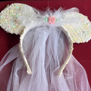 May include: A white sequined mouse ear headband with a tulle veil. The ears are covered in iridescent sequins. A white bow with feathers and a pink rose decorates the top. The veil is attached to the headband.