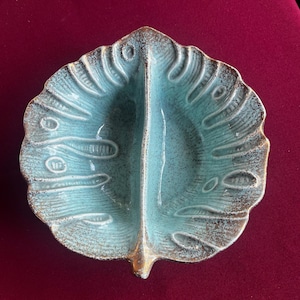 Puede incluir: Un plato de cerámica turquesa con forma de hoja y diseño dividido. El plato tiene una superficie texturizada con aspecto moteado y un borde dorado. El diseño de la hoja presenta vetas detalladas y un borde decorativo.
