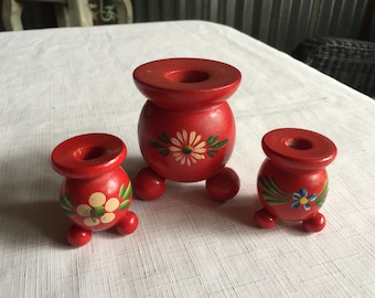 May include: Three red wooden candle holders with painted floral designs. The largest holder has a white daisy with green leaves, the smaller holders have a white flower with green leaves and a blue flower with yellow center.