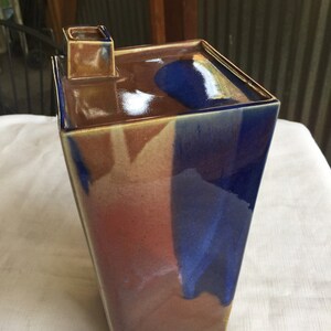 Op de afbeelding: Een vierkante keramische vaas met een unieke glazuur in tinten bruin, blauw en wit. De vaas heeft een klein tuitje aan de bovenkant.