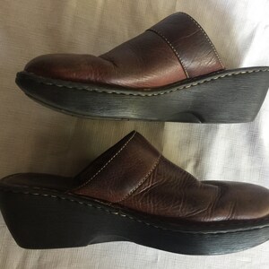 May include: A pair of brown leather clogs with a thick platform sole. The clogs have a strap across the top of the foot and a closed toe.