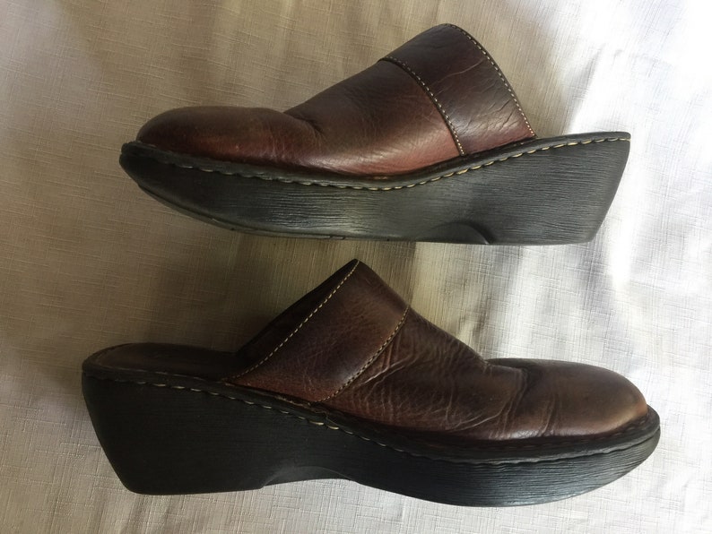 May include: A pair of brown leather clogs with a thick platform sole. The clogs have a strap across the top of the foot and a closed toe.
