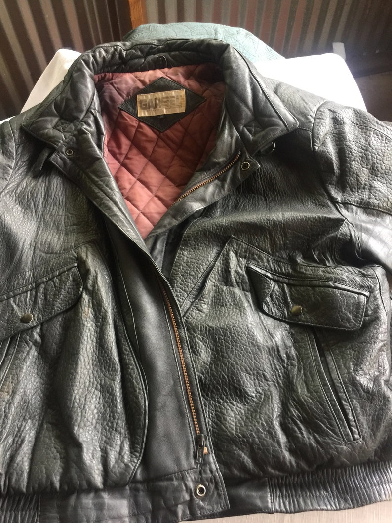 Pu&ograve; includere: Una giacca bomber in pelle nera con chiusura a zip e fodera trapuntata. La giacca ha un'etichetta che dice "G.A.R.S.E.L."