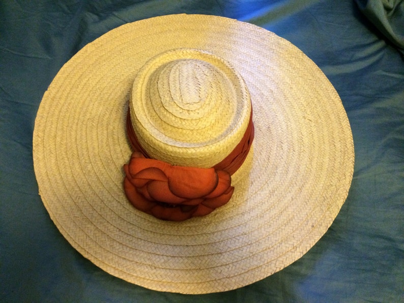 Pu&ograve; includere: Un grande cappello di paglia a tesa larga con un nastro rosso e bianco attorno alla corona. Il cappello &egrave; decorato con un grande fiore rosso.