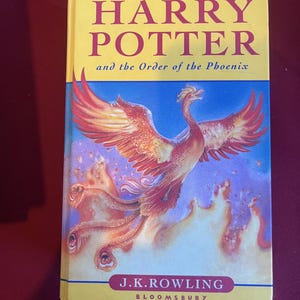 May include: A hardback book titled Harry Potter and the Order of the Phoenix by J.K. Rowling, published by Bloomsbury. The cover features a large, vibrant orange and red phoenix rising from flames against a blue background with yellow borders.