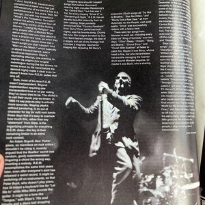May include: Black and white photo of a man standing on a stage with a microphone in his hand. He is wearing a light-colored shirt and dark pants. The stage is lit with spotlights. The photo is from a concert.