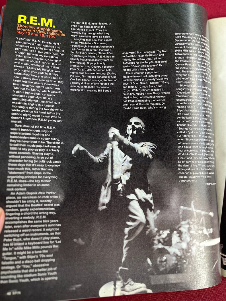 May include: Black and white photo of a man standing on a stage with a microphone in his hand. He is wearing a light-colored shirt and dark pants. The stage is lit with spotlights. The photo is from a concert.